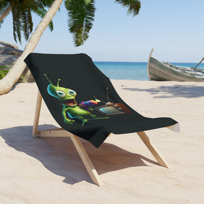 Black premium beach towel with a colorful 3D alien holding a retro TV remote, displayed on a wooden lounge chair at a tropical beach.