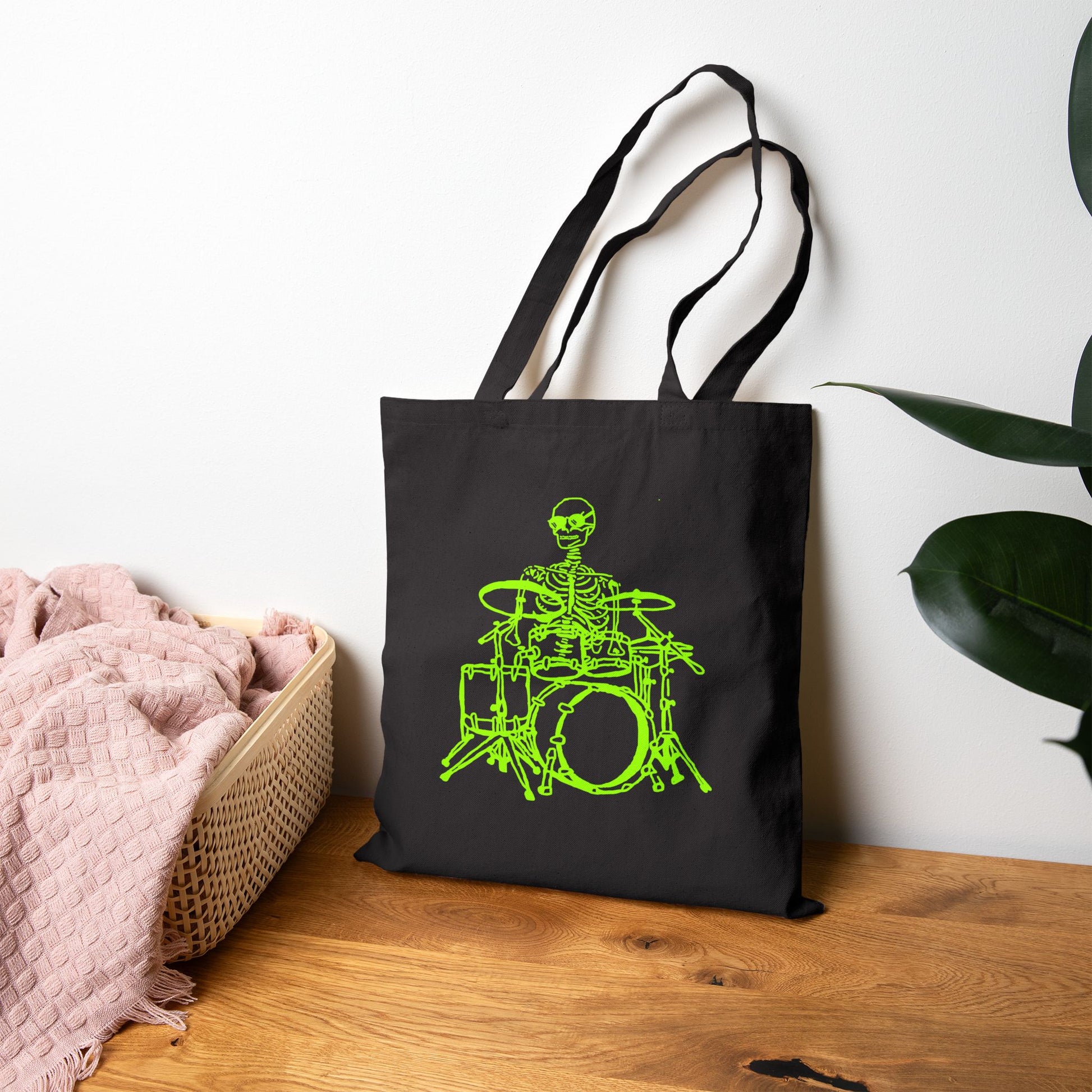 Black tote bag with a green skeleton playing drums design on a wooden surface.