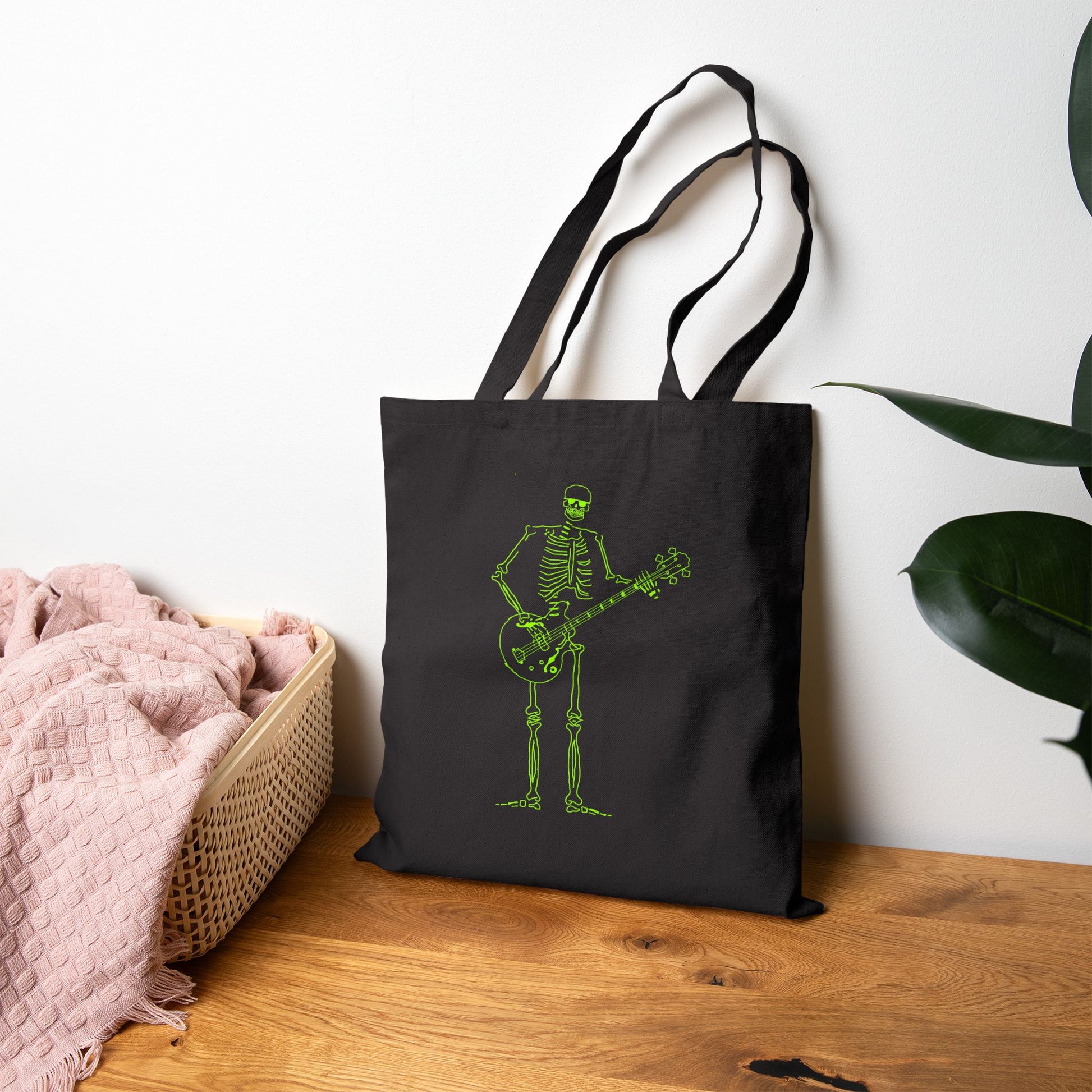 Black tote bag with a unique green skeleton playing guitar design on a wooden surface.