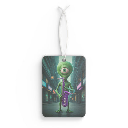 Rectangular car air freshener showing a green, one-eyed cyclops alien holding a purple saxophone on a dark city street under neon lights.