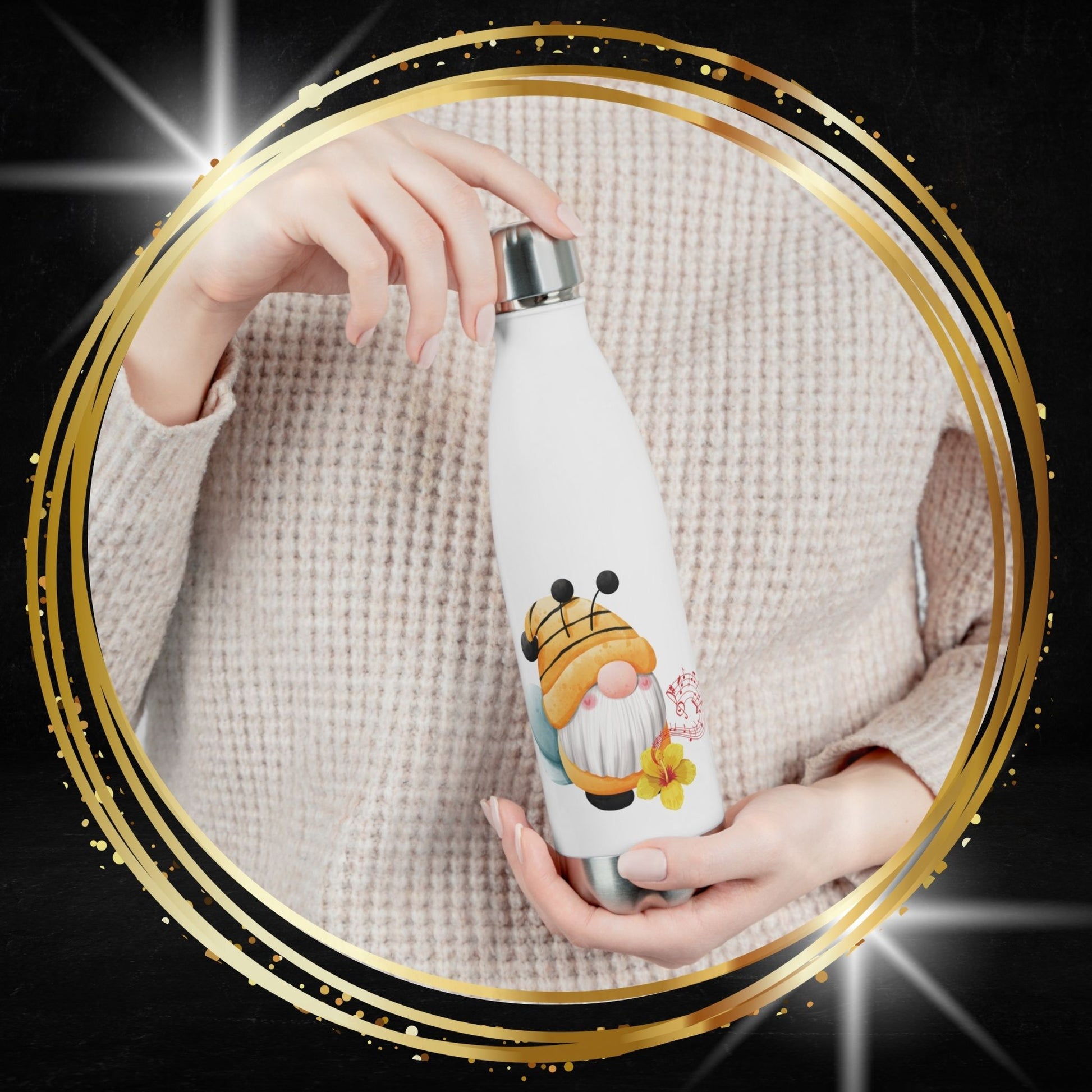 A white insulated water bottle with a silver cap, decorated with a cute cartoon bee gnome character, held by hands wearing a beige knit sweater. 