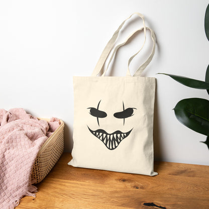 Beige tote bag with a halloween black face design on a wooden surface with a white wall and plant in the background.