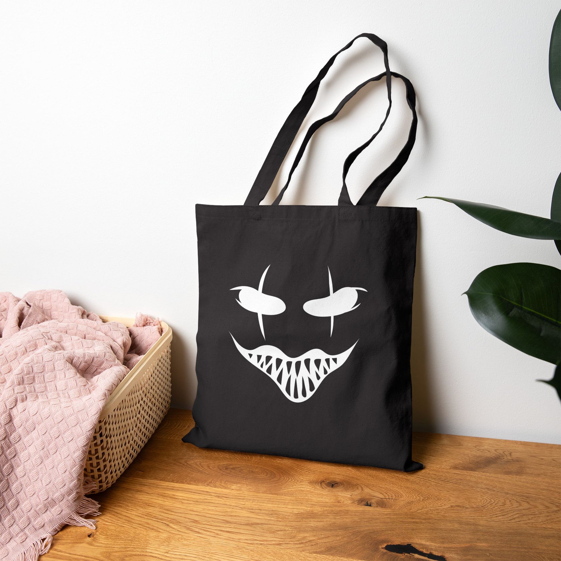 Black tote bag with a white face design on a wooden surface with a plant in the background