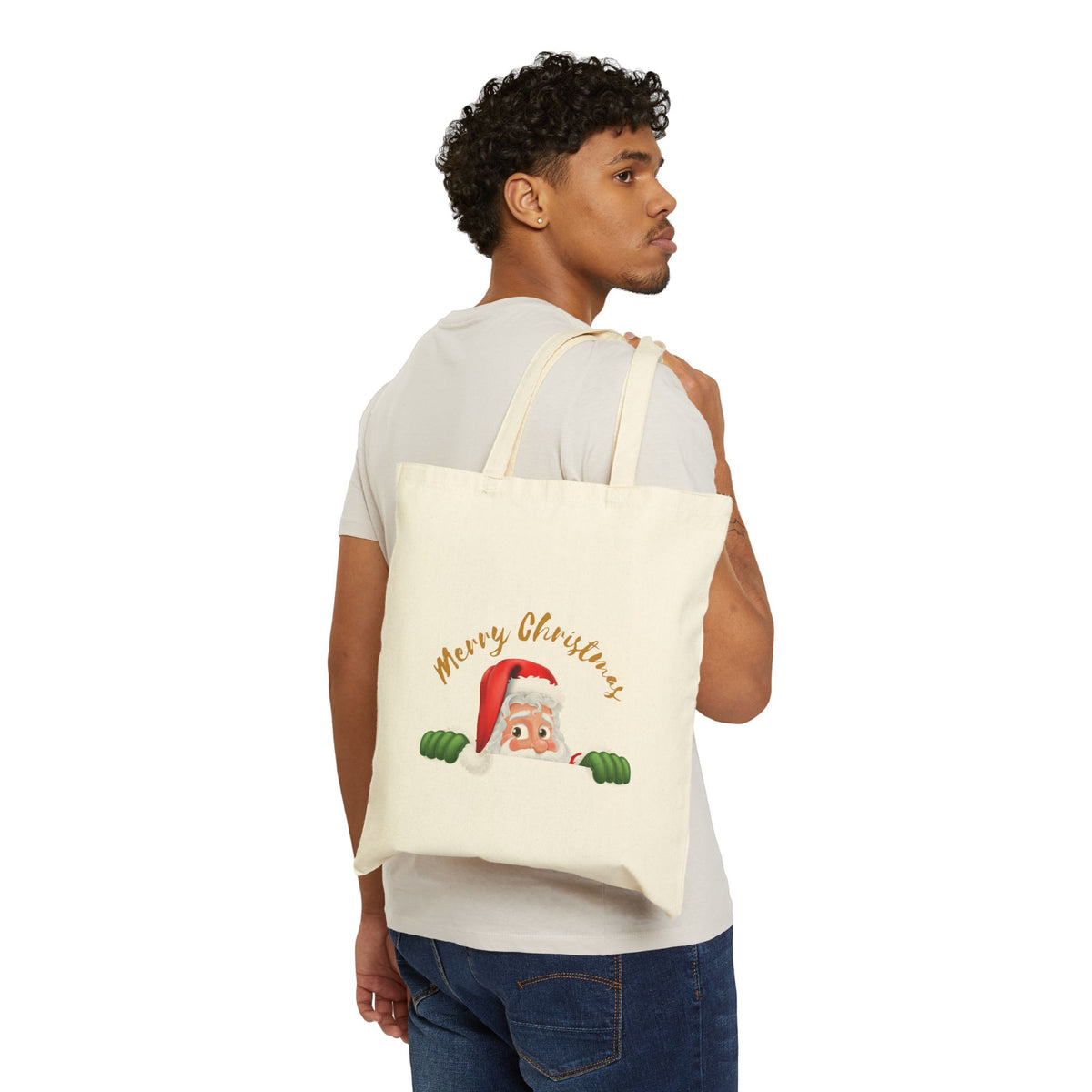 Man carrying a beige cotton Christmas tote bag over his shoulder printed with a cute peeking Santa hat artwork and holiday typography.   