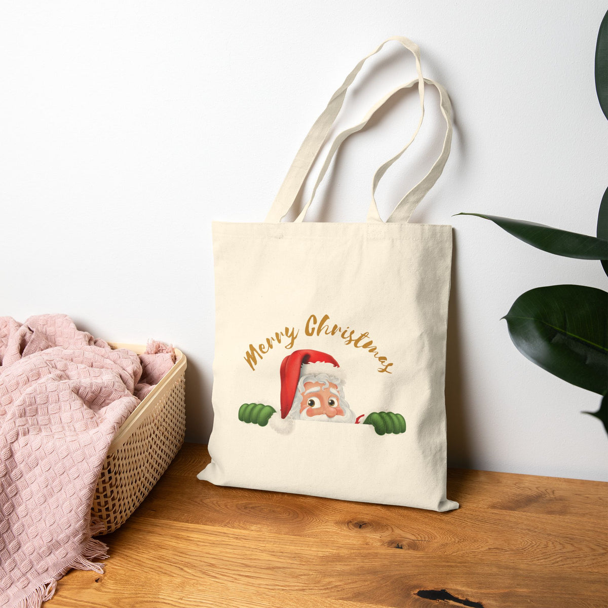 Natural canvas tote bag leaning on a wooden table featuring a cute peeking Santa Claus design with gold Merry Christmas lettering for holiday shopping.   