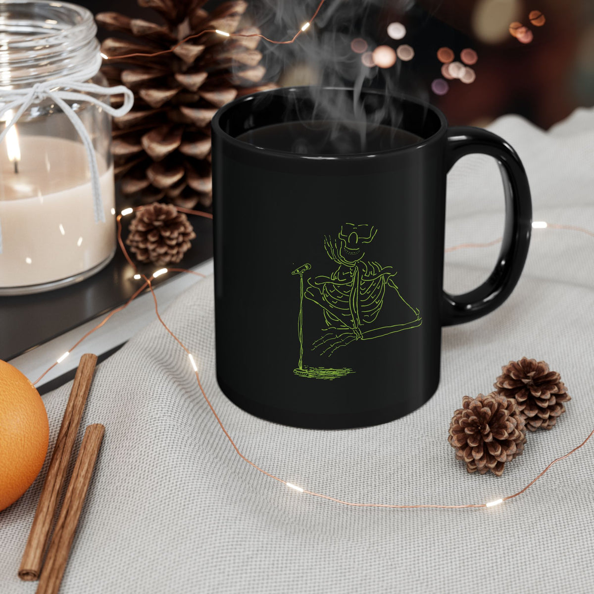 Steaming coffee in a black ceramic mug with a fluorescent green skeleton playing drums illustration on a warm wooden table surrounded by pinecones and candles.