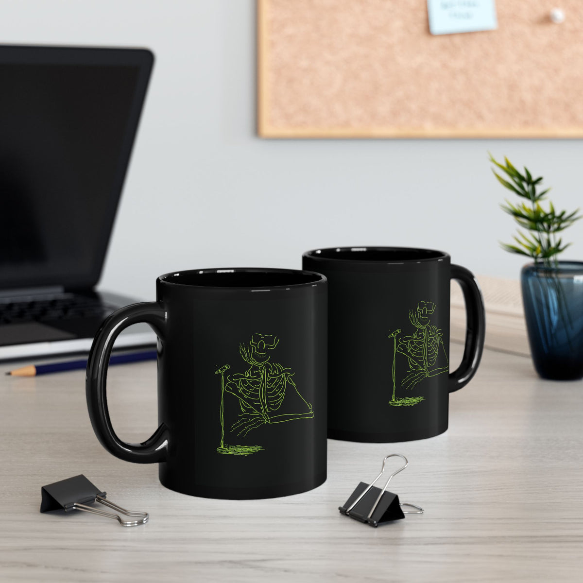 Pair of black ceramic coffee cups featuring glowing neon green skeleton musicians sitting on a modern office desk next to a laptop and binder clips.