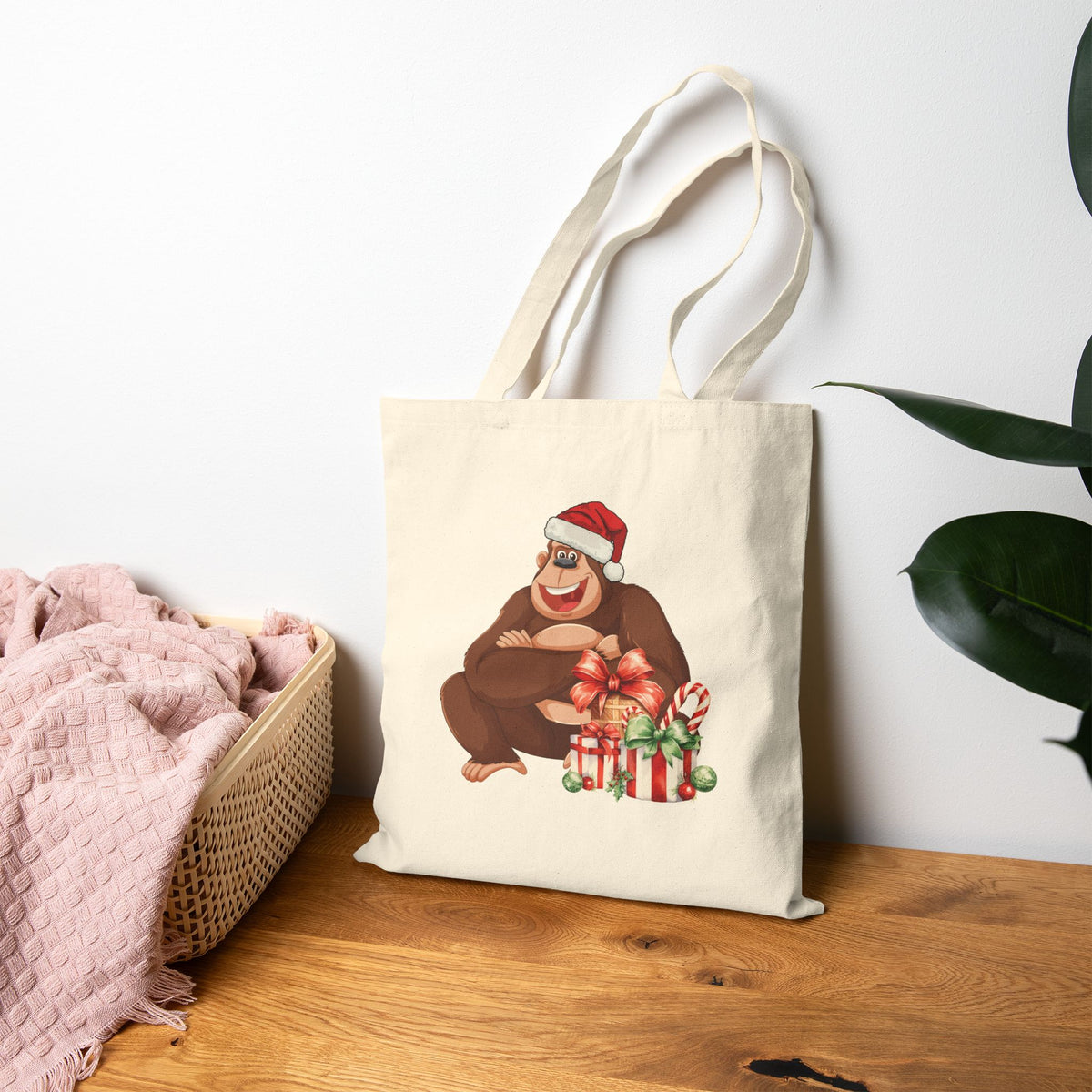 Natural canvas tote bag leaning on a wooden table featuring a smiling gorilla wearing a Santa hat next to wrapped holiday gifts and candy canes for festive shopping.   